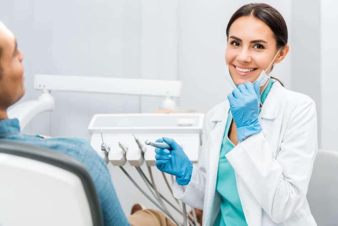 cheerful female dentist holding drill and smiling near patient